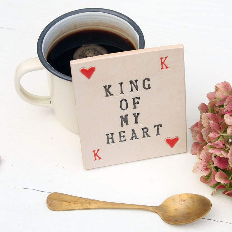 small white ceramic rectangle coaster with black text king of my heart
in left top corner and bottom right corners are red hearts and red k in the other corners