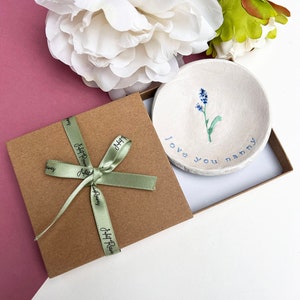 May include: A small white ceramic dish with a blue flower design and the text "love you nanny". The dish is in a brown gift box with a green ribbon.