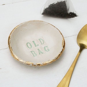 Small round ceramic dish, finished in off white glaze. The text old bag printed in the centre and hand painted in sage green. Gold leaf applied to the rim of the dish. Gold teaspoon.