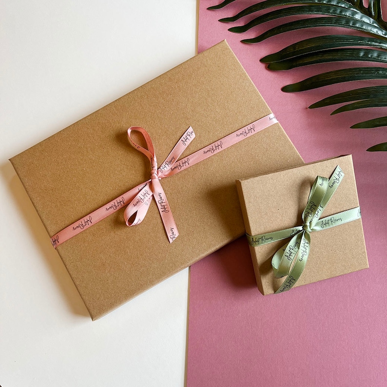 May include: Two brown gift boxes with pink and green ribbon tied in bows. The boxes are on a pink and white surface. The ribbon on the larger box has the text "Happy Place" printed on it.