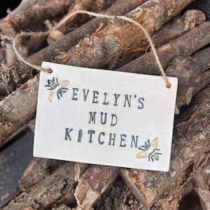 May include: A white rectangular sign with the words "EVELYN'S MUD KITCHEN" in black. The sign features bee illustrations and is suspended by twine. The background consists of a pile of brown twigs.