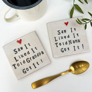 K&ouml;nnte beinhalten: Zwei wei&szlig;e Keramik-Untersetzer mit schwarzem Text und einem roten Herzen. Der Text auf den Untersetzern lautet "Saw It Liked It Told Grandpa Got It!" und "Saw It Liked It Told Nana Got It!"