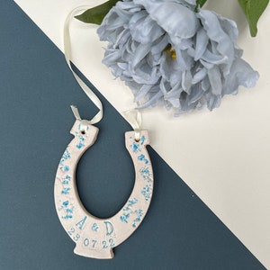 White ceramic glazed horseshoe with pretty blue shades of flowers up the sides, with text: INITIAL & INITIAL DATE. Text and date along the bottom. Cream ribbon attached to the top for hanging purposes.