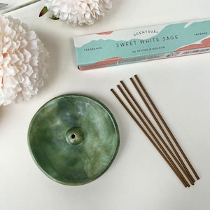 May include: A green ceramic incense holder with a small hole in the centre.  A box of incense sticks labelled "Scentual Sweet White Sage 30 Sticks & Holder" is in the background.  The incense sticks are brown and are arranged in a bundle.