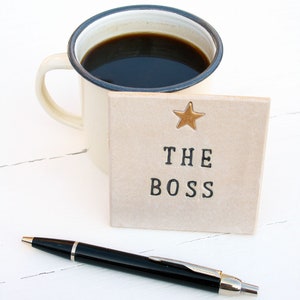 A square white coaster with a gold painted star at the top and black text: THE BOSS