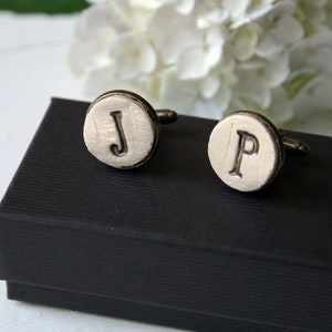 May include: A pair of cufflinks in a black box. The cufflinks are round and white with the letters "J" and "P" engraved on them.