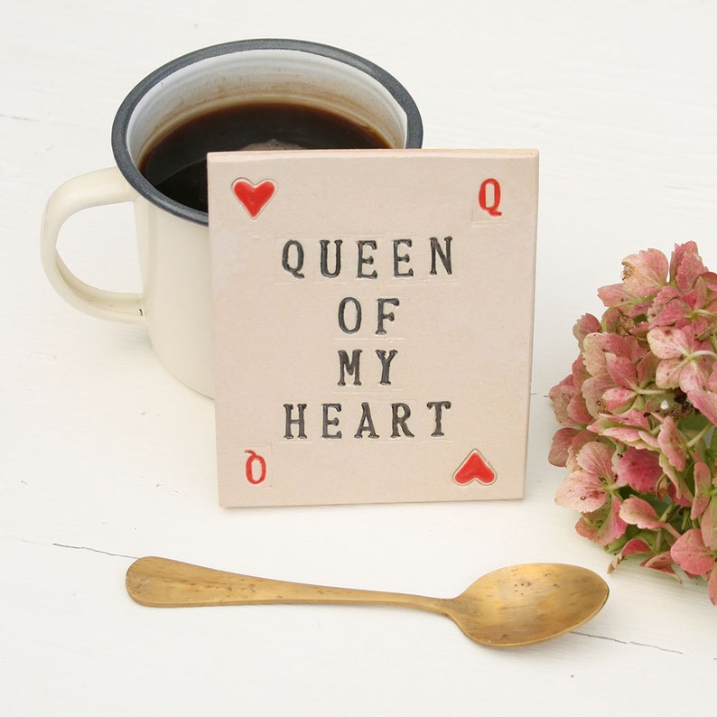 small white ceramic rectangle coaster with black text queen of my heart
in left top corner and bottom right corners are red hearts and red Q in the other corners