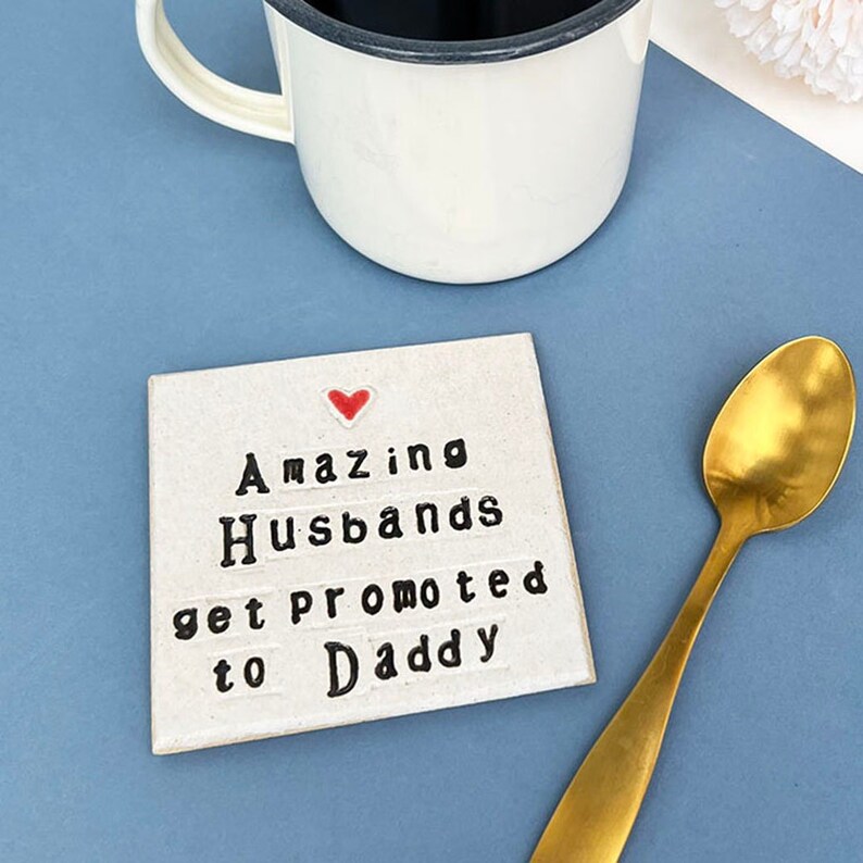 Square ceramic coaster finished in an off white glaze. Black text reads, amazing husbands get promoted to daddy. small red heart above the text. Gold teaspoon.