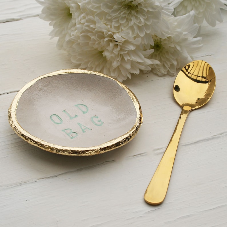 Small round ceramic dish, finished in off white glaze. The text old bag printed in the centre and hand painted in sage green. Gold leaf applied to the rim of the dish. Gold teaspoon.