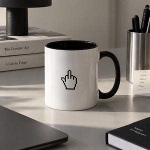 Può includere: Una tazza in ceramica bianca con interno e manico neri. L'esterno presenta una grafica nera pixelata di una mano con il dito medio alzato, che ricorda l'icona di un cursore del computer. La tazza ha una forma cilindrica e liscia.