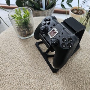 Puede incluir: Un mando de videojuegos negro apoyado sobre un soporte de plástico negro impreso en 3D. El mando cuenta con dos joysticks analógicos, una cruceta, cuatro botones de acción y un pequeño trozo de cinta blanca con una marca cuadrada marrón.