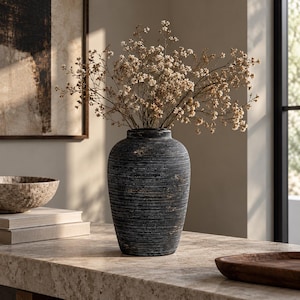 May include: A dark charcoal gray vase with a textured, horizontally ribbed surface and a weathered finish. The vase has a rounded body and a narrow neck, and it is filled with a bouquet of delicate, dried off-white baby's breath flowers.