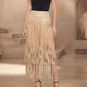 May include: A sleeveless black mock neck top paired with a tan midi skirt featuring a smooth waistband and two tiers of long, flowing fringe. The outfit is completed with black strappy high-heeled sandals, creating a coordinated ensemble.