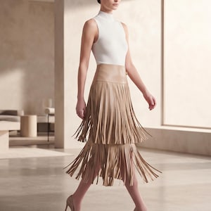 May include: A sleeveless white mock neck top paired with a tan midi skirt featuring two tiers of long, vertical fringe detailing. The outfit is completed with matching tan pointed-toe high-heeled pumps, creating a coordinated monochromatic look.