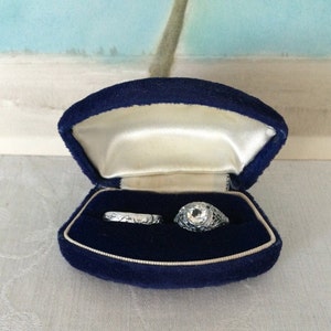 May include: A silver-toned ring with a central clear stone and intricate filigree metalwork alongside a matching silver-toned band with an engraved pattern, both presented inside a dark blue velvet jewelry box with a cream-colored satin lining.