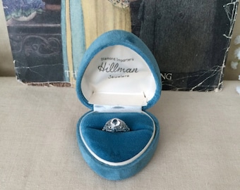 Puede incluir: Un anillo de tono plateado con una piedra central transparente y un intrincado trabajo de metal en filigrana, presentado dentro de un joyero de terciopelo azul en forma de corazón. La tapa interior tiene el texto: Diamond Importers Hillman Jewelers.