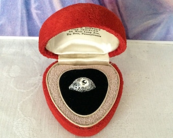 May include: A silver-toned filigree ring with a central stone sits inside a heart-shaped red velvet jewelry box. The interior lid features printed text: John R. Shively, The Jeweler, 34 West Long Ave., Du Bois, Pennsylvania.