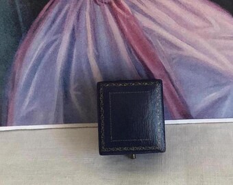 Puede incluir: Un joyero cuadrado de color azul oscuro con una superficie texturizada y bordes decorativos dorados en filigrana. La caja presenta un marco rectangular central en relieve en la tapa y una pequeña bisagra metálica visible en la parte inferior.