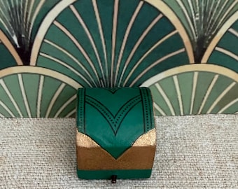May include: A small, rectangular box featuring a vibrant green finish with metallic gold accents on the lower corners. The top surface displays a dark, intricate V-shaped pattern with fine dotted lines, and a small metal latch is visible at the base.