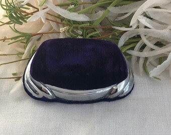 May include: A small, rectangular jewelry box featuring a deep purple velvet top and a polished silver-tone metal base. The metal trim has a smooth, curved design along the front edge, providing a contrast to the soft, dark fabric of the lid.