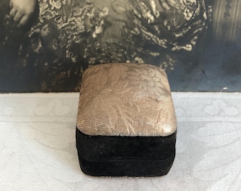 May include: A rectangular jewelry box featuring a textured, light beige lid with a subtle floral pattern and a solid black velvet base. The box is closed and has a structured, compact shape with a flat top surface and straight, defined edges.