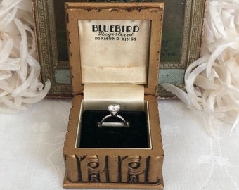 Puede incluir: Un anillo de tono plateado con una piedra de corte redondo en un engaste de garras, exhibido dentro de una caja dorada. La tapa interior de la caja presenta el texto BLUEBIRD REGISTERED DIAMOND RINGS impreso en negro sobre tela crema.