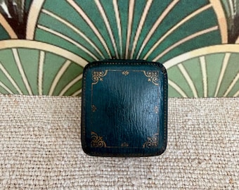 May include: A small, square, dark teal jewelry box with rounded corners. The lid features a gold-colored embossed border with decorative filigree patterns at each corner. The surface of the box has a subtle, textured, leather-like appearance.