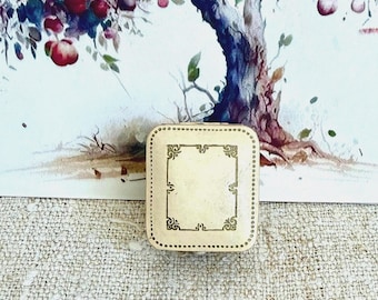 May include: A small, square, cream-colored jewelry box with rounded corners and a decorative gold-toned border pattern on the lid. The box features a smooth, solid surface with a symmetrical, ornate frame design centered on the top cover.