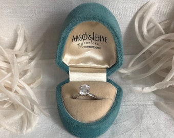 Puede incluir: Un anillo plateado con una piedra redonda transparente en un joyero de terciopelo azul verdoso. El interior de la tapa presenta el texto impreso Argo & Lehne Jewelers Columbus, Ohio, con un forro de satén color crema e inserto de terciopelo.