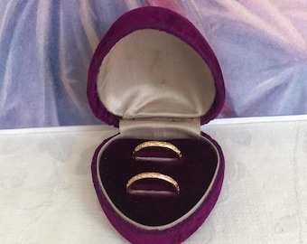 Puede incluir: Dos anillos finos de oro con un patrón grabado descansan dentro de un joyero de terciopelo morado oscuro en forma de corazón. El interior del joyero está forrado con una tela de satén beige claro, contrastando con el exterior de terciopelo.