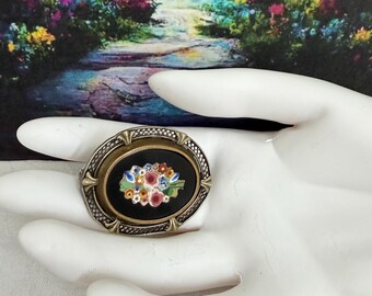 May include: An oval brooch featuring a central black inlay decorated with a colorful floral micro-mosaic pattern. The piece is framed by a textured, metallic border with decorative prongs at the top, bottom, and sides, creating a detailed accessory.