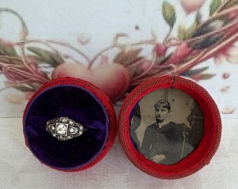 May include: A silver-toned ring with a central stone and smaller accent stones, nestled in a round red box with a purple velvet interior. The box lid features a circular black and white photograph of a person wearing a high-collared garment.