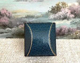 May include: A square, dark teal jewelry box with a textured, pebbled leather-like finish. The lid features two curved, gold-colored stitched lines that frame the center, creating a distinct geometric pattern on the top surface of the container.