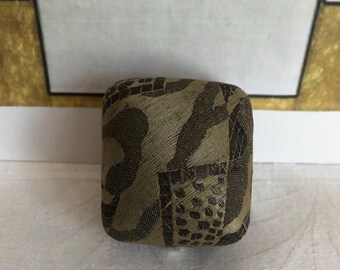 May include: A small, square-shaped fabric-covered object featuring a textured, woven pattern in shades of olive green and dark brown. The surface displays a mix of abstract shapes and a grid-like dotted design, with rounded corners and a firm structure.