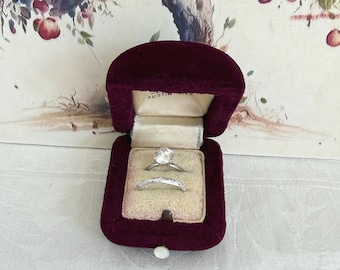 May include: A deep burgundy velvet ring box with a cream-colored interior lining, containing two silver-toned rings. The top ring features a single round clear stone, while the bottom ring has a textured, patterned band. The box is open.