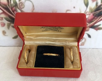May include: A set of three gold-toned rings with engraved patterns, presented in a rectangular red jewelry box with a cream-colored interior lining and black velvet ring slots. The box lid features the text "Kosch's, Inc. JEWELERS SPARTANBURG, S.C."
