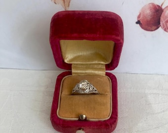May include: A silver-toned ring featuring a central round-cut stone set within an intricate, filigree-style metal band. The ring is displayed inside an open, rectangular, deep red velvet jewelry box with a gold-colored satin interior lining.