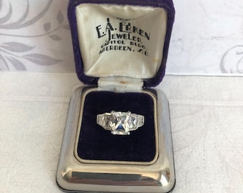 May include: A silver-toned ring with a large rectangular center stone and smaller baguette-cut side stones, presented in a purple velvet-lined jewelry box. The box lid features the text E.A. ERKEN JEWELER CAPITOL BLDG. ABERDEEN, S.D.