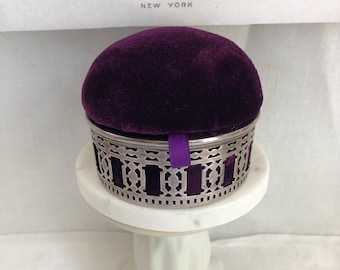 May include: A round pincushion featuring a deep purple velvet top and a silver-toned metal base with an intricate lattice cutout design. A small purple ribbon tab extends from the side of the silver frame. The item sits atop a white pedestal.