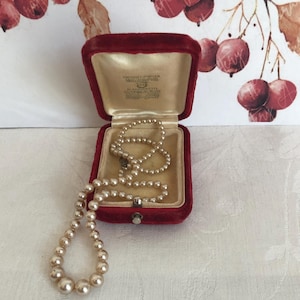May include: A string of cream-colored pearls with a graduated size design, resting inside an open, deep red velvet jewelry box. The interior of the box features a cream-colored satin lining with printed text that reads K. MIKIMOTO.