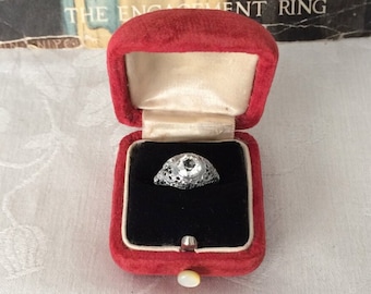 May include: A silver-toned ring with a central clear stone and intricate filigree metalwork, presented inside a red velvet-lined box with a cream-colored interior. The box features a small white button closure and a cream-colored base.