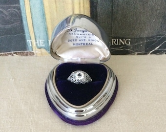 May include: A silver-toned heart-shaped ring box with a deep purple velvet interior containing a filigree ring with a clear center stone. The interior lid features the text D. Jacques Diamantaire Suite 5 2022 Ave. Union Montreal in black ink.