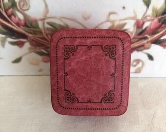 May include: A square-shaped, textured red box with rounded corners. The top surface features a dark, intricate, decorative border design consisting of a dotted line framing a central rectangular area with ornate, swirling corner flourishes.