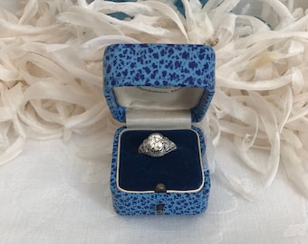 Puede incluir: Un anillo de tono plateado con una piedra central de corte redondo y filigrana metálica intrincada, presentado dentro de un pequeño joyero cuadrado moteado en azul y negro con forro interior de terciopelo azul oscuro y tapa.