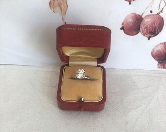 Puede incluir: Un anillo de tono plateado con una piedra redonda transparente engastada en garras. El anillo se presenta dentro de un pequeño joyero rectangular de textura de cuero rojo oscuro, con un forro de terciopelo beige y una tapa abatible.