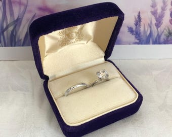 Puede incluir: Un anillo de compromiso plateado con una piedra redonda facetada y una alianza a juego con un patrón grabado, ambos dentro de un joyero de terciopelo morado oscuro con forro interior color crema y un logotipo dorado.