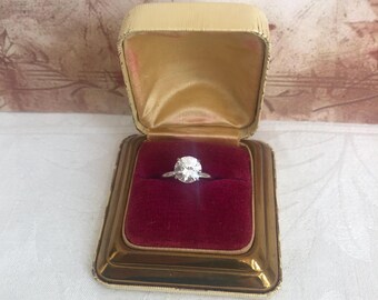 Puede incluir: Un anillo de tono plateado con una piedra redonda transparente y facetada en un engaste de garras. El anillo se presenta dentro de un joyero rectangular con bisagras, exterior dorado metálico y un forro de terciopelo rojo intenso.