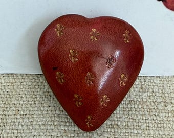 May include: A heart-shaped box with a smooth, deep red leather exterior. The surface is decorated with a repeating pattern of small, gold-colored embossed lion motifs scattered across the front. The object has a rounded, puffed silhouette.
