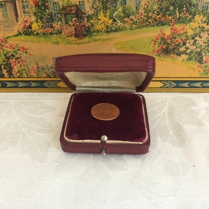May include: A copper penny displayed inside a small, burgundy velvet-lined jewelry box. The box is open, revealing the coin against the plush interior. The box has a clasp closure and a cream-colored satin lining.