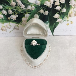 May include: A diamond ring in a white, heart-shaped jewelry box with a green velvet interior. The ring is set against a backdrop of a floral illustration featuring white flowers and green leaves. The box is open, showcasing the ring.